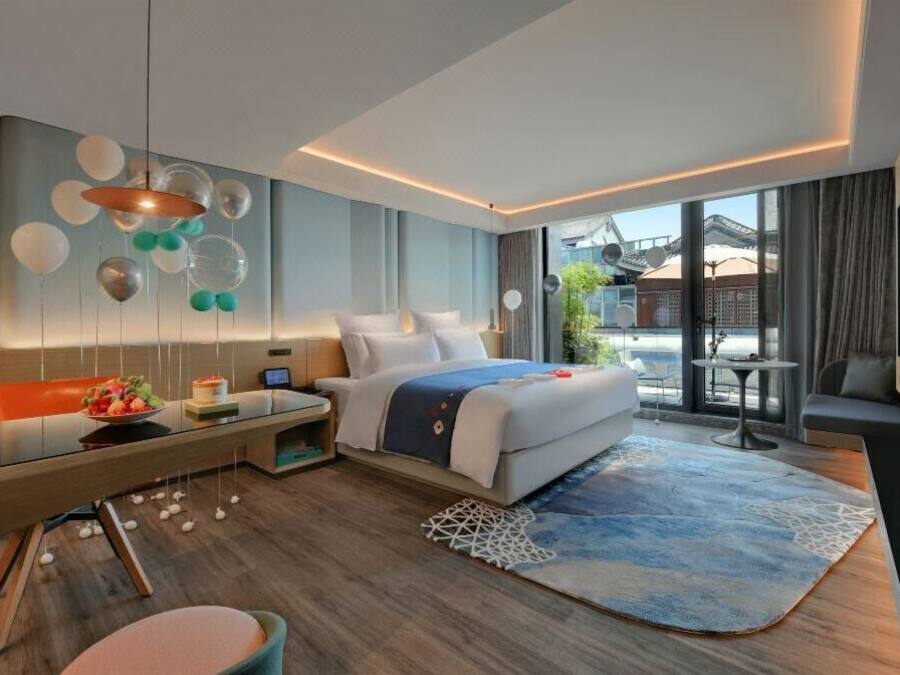 A modern hotel room with a large bed, a desk, a dining table, and a view of the outside.