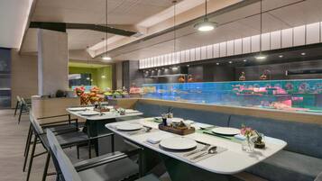 A modern restaurant with long communal tables, a central bar area, and a vibrant aquarium display.