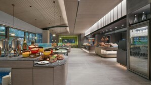 Un buffet de petit-déjeuner moderne proposant une variété d’aliments et de boissons, un espace repas avec des tables et une grande fenêtre laissant entrer la lumière naturelle.