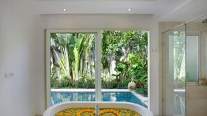 Un baño moderno con una bañera independiente llena de agua y flores amarillas, un box de ducha de vidrio, y vistas a una piscina y abundante vegetación exterior.