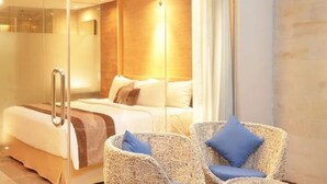 Una habitación de hotel moderna con cabina de ducha de vidrio, una cama con cubrecama blanco y dos sillas de mimbre con cojines azules.