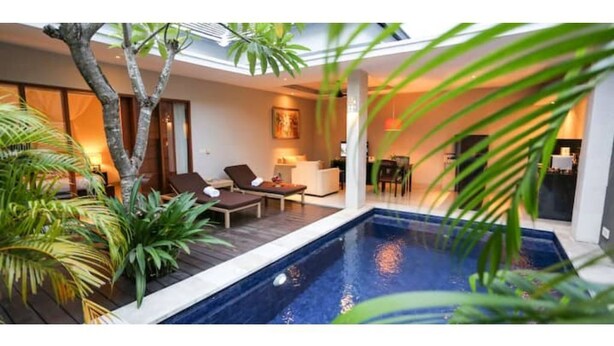 A modern indoor pool area with lounge chairs, a small table, and a dining area visible in the background.