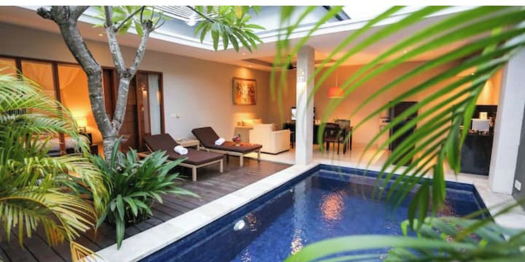 Un espace piscine intérieur moderne, avec des chaises longues, un coin repas et une cuisine en arrière-plan.