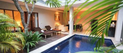 Un espace piscine intérieur moderne, avec des chaises longues, un coin repas et une cuisine en arrière-plan.