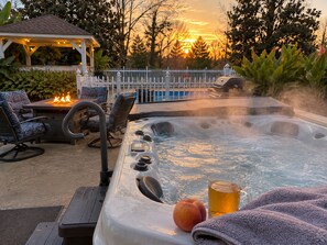 A hot tub with a peach and a glass of amber liquid, surrounded by outdoor furniture and a pool.