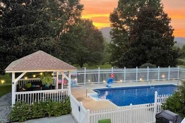 Una piscina con un deck cercado, un pabellón y vistas a árboles y un atardecer.