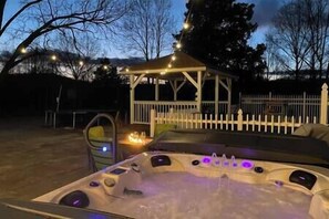 Un jacuzzi con cubierta de madera y luces colgantes, rodeado por un cerco blanco y árboles.