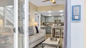 A modern living room with a sofa, coffee table, and a glass door leading to a kitchen area with cabinets and a dining table.