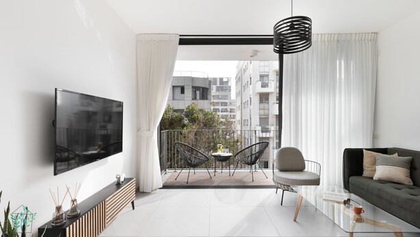 Een moderne woonkamer met een flatscreen televisie, een bank, een eettafel met stoelen, en een balkon met uitzicht op gebouwen.