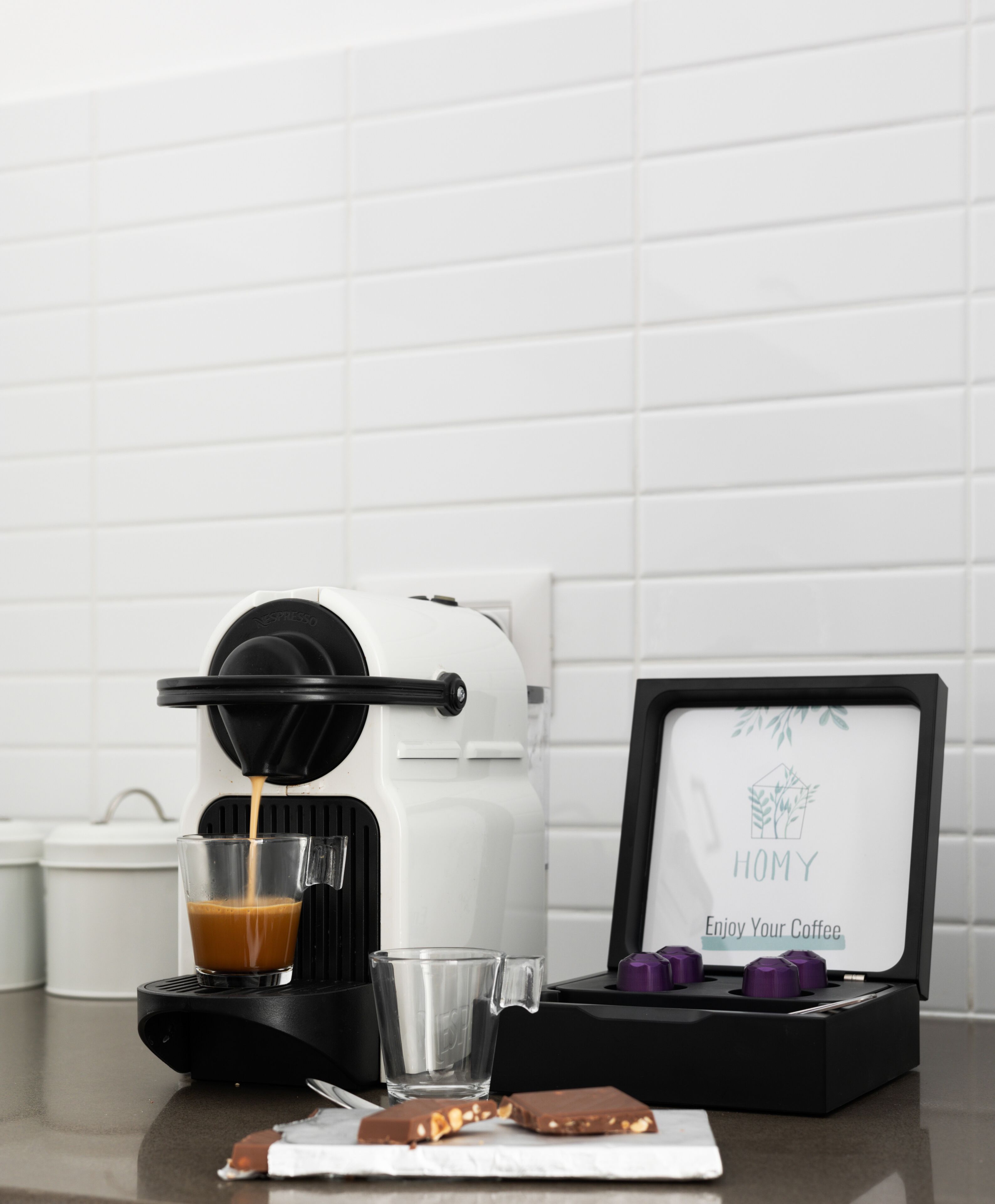 A coffee machine dispensing coffee into a glass, a box with a "HOMY" label, and a chocolate bar on a countertop.