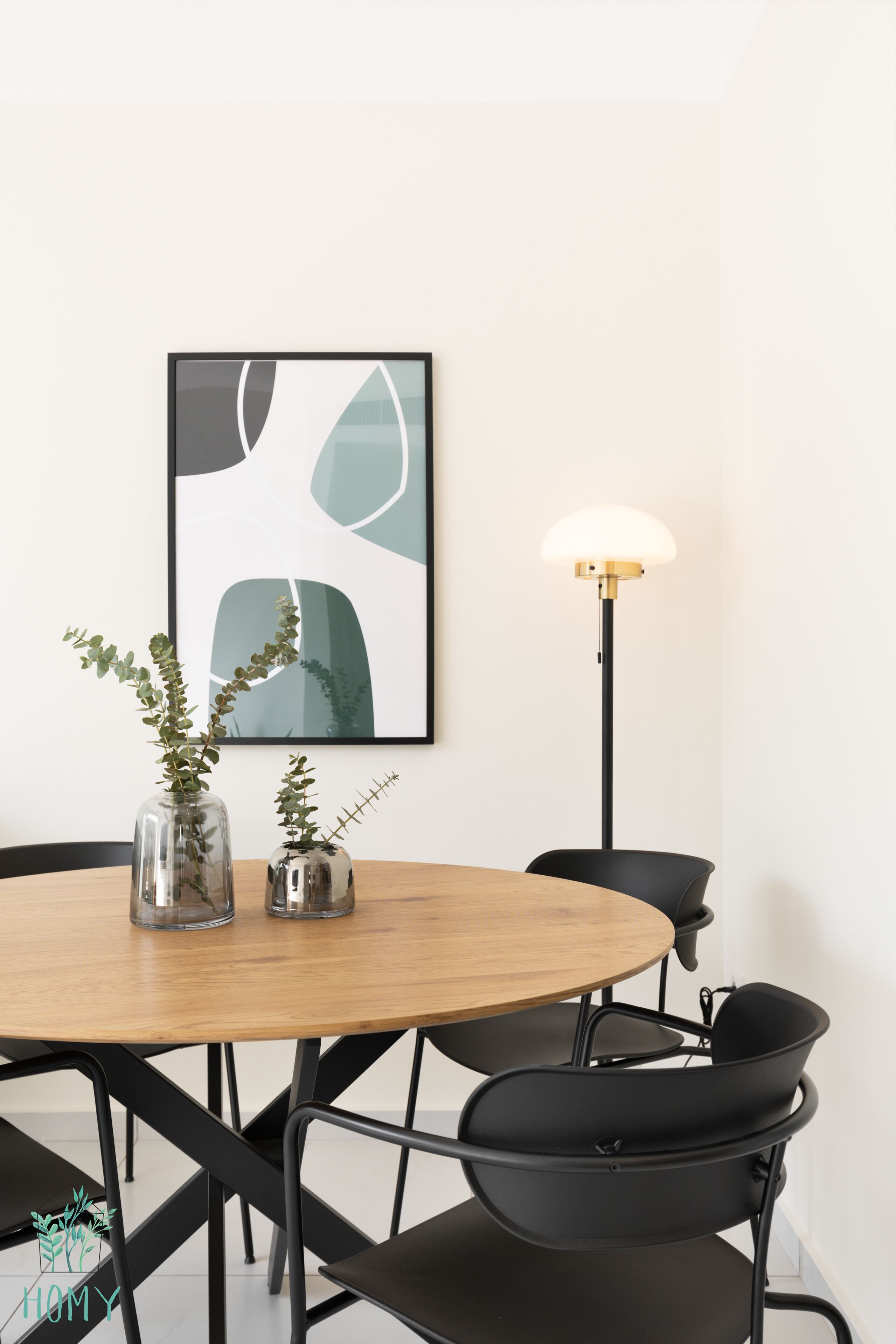 A dining area with a round wooden table, black chairs, two glass vases with plants, a modern floor lamp, and a framed abstract artwork on the wall.