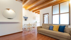 Una sala de estar moderna con techo de madera, un sofá beige con cojines azules y amarillos, un espejo redondo en la pared y una zona de comedor al fondo.