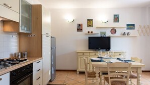 Una cocina moderna con armarios blancos, estufa, horno y refrigerador. Hay una mesa de comedor con sillas y un televisor de pantalla plana sobre un mueble bajo.