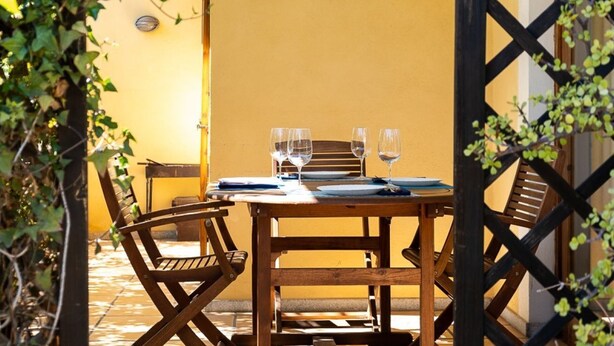 Una mesa de madera con tres copas de vino y un menú, rodeada de sillas de madera, frente a una pared amarilla con celosía.