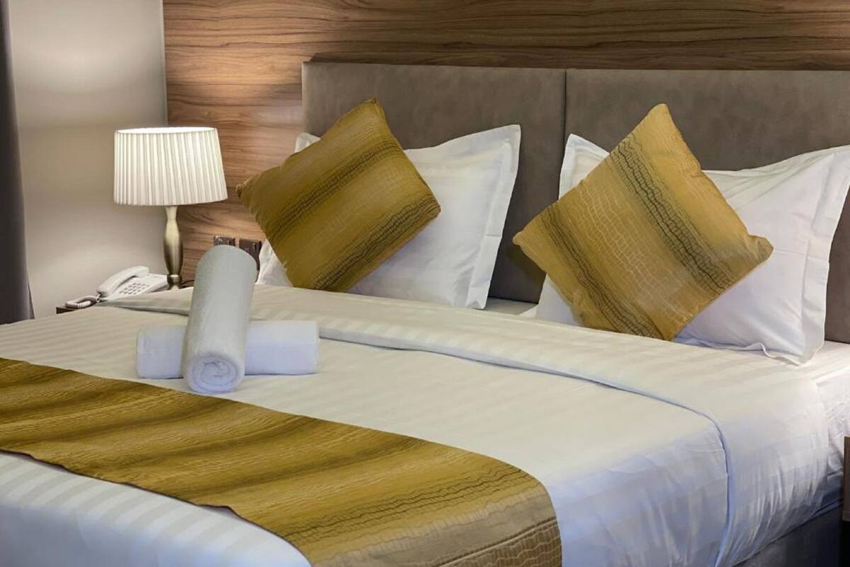 A neatly made bed with a white and gold striped blanket, two gold pillows, and a white pillow. A white lamp and a rolled white towel are on the bed.