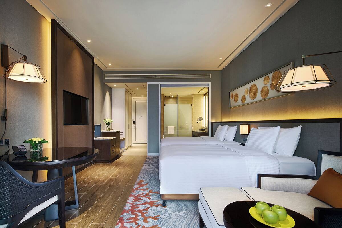 Habitación de hotel moderna con una cama grande, un escritorio con una silla, una mesita de noche con lámpara y un baño visible a través de una puerta abierta.