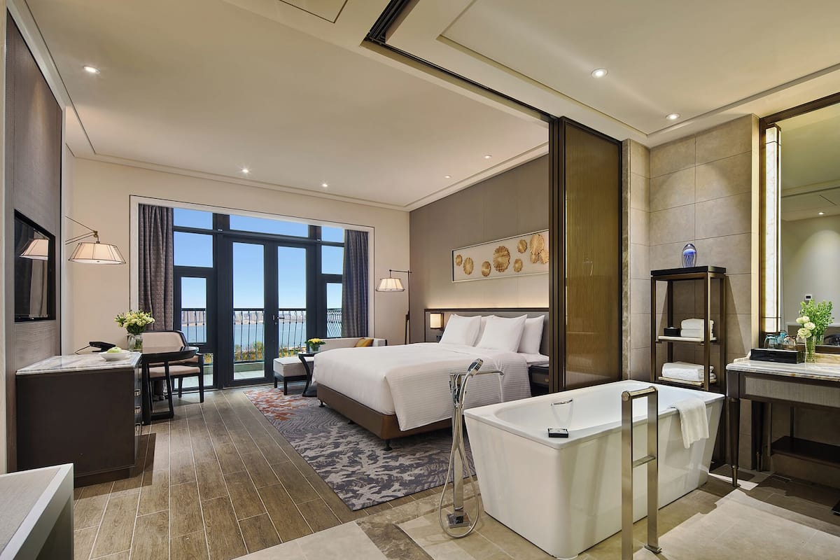 Habitación de hotel moderna con una cama grande, baño independiente con bañera, escritorio con silla y vistas al exterior a través de amplias ventanas.