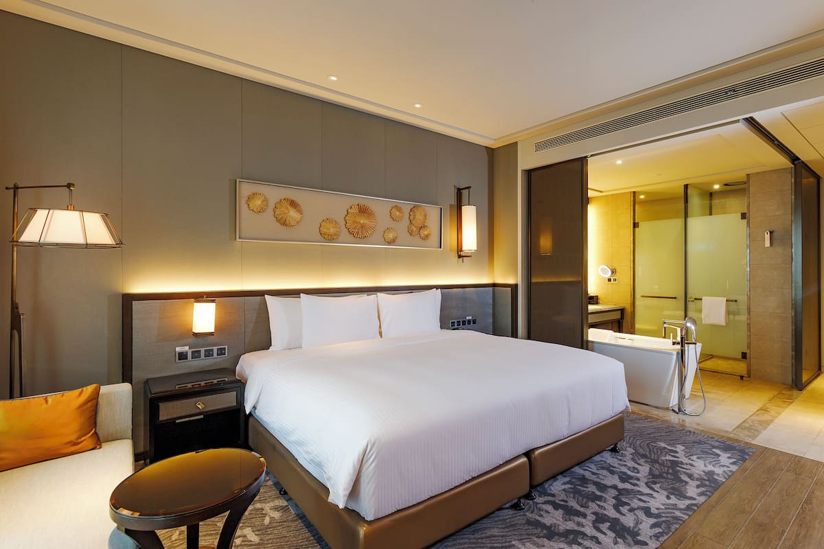 Habitación de hotel con una cama grande, una mesita de noche, una lámpara, una obra de arte en la pared y un baño visible a través de una puerta abierta.