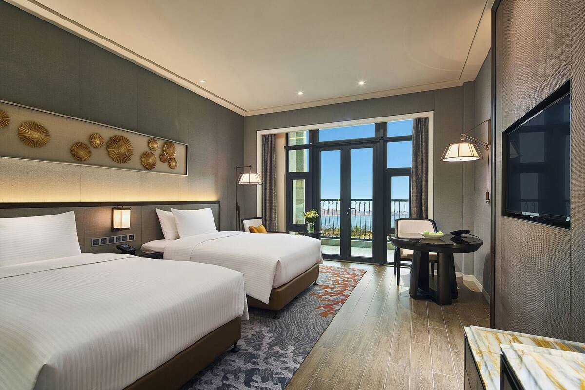 Habitación de hotel con dos camas, un escritorio, televisión y balcón con vistas.