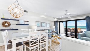 Un salon moderne avec un coin repas, de grandes fenêtres offrant une vue sur l’océan et un ventilateur de plafond.