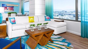 A modern living room with a white sofa, blue and yellow cushions, a wooden coffee table, and a view of the city through large windows.