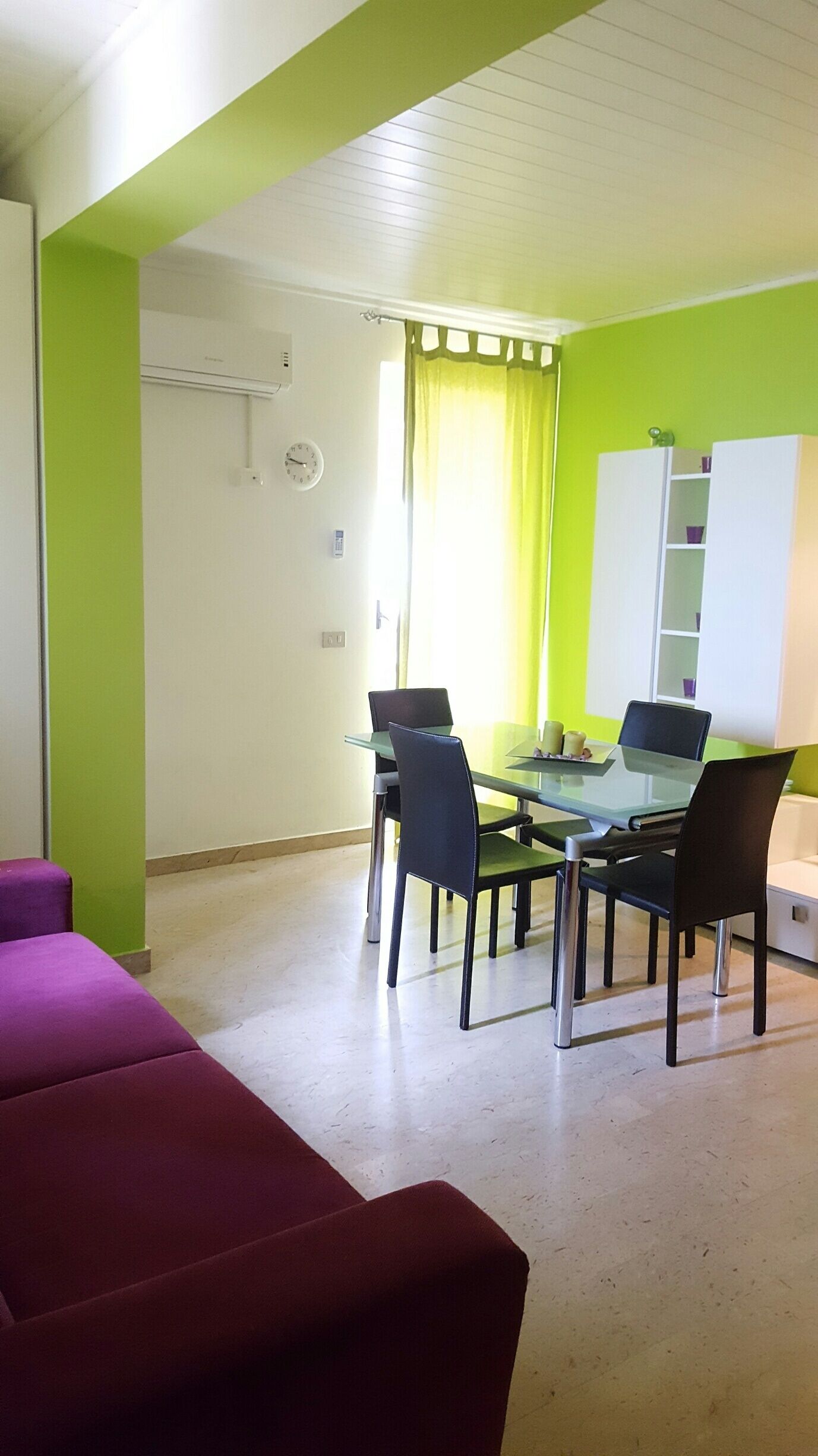 A dining area with a glass table and chairs, a clock on the wall, and a green accent wall.