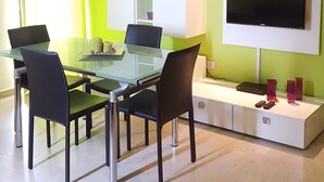 A modern living room with a glass dining table, four chairs, a flat-screen TV mounted on the wall, and a white cabinet with shelves. - Residence Eloisa  (Balestrate)