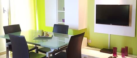 A modern living room with a glass dining table, four chairs, a flat-screen TV mounted on the wall, and a white cabinet with shelves.