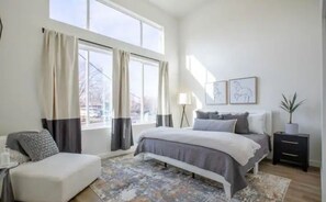 Relax in this serene bedroom with a large bed, armchair, and potted plant, surrounded by framed artworks and a patterned rug.