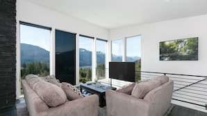 Ein modernes Wohnzimmer mit einer Sofagarnitur, einem Couchtisch und einem Fernseher. Große Fenster bieten Blick auf die Berge.