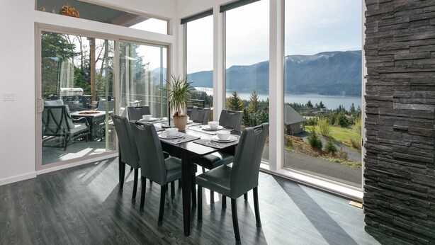 Ein moderner Essbereich mit einem großen Tisch, Stühlen und Blick auf einen See und Berge.