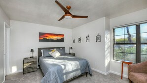 Ein Schlafzimmer mit Bett, Nachttischen, Deckenventilator, Fenster mit Blick und Wandbildern.