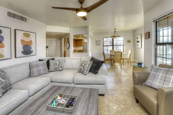 A spacious living room with a grey sectional sofa, a coffee table, and a dining area with wicker chairs and a glass table.