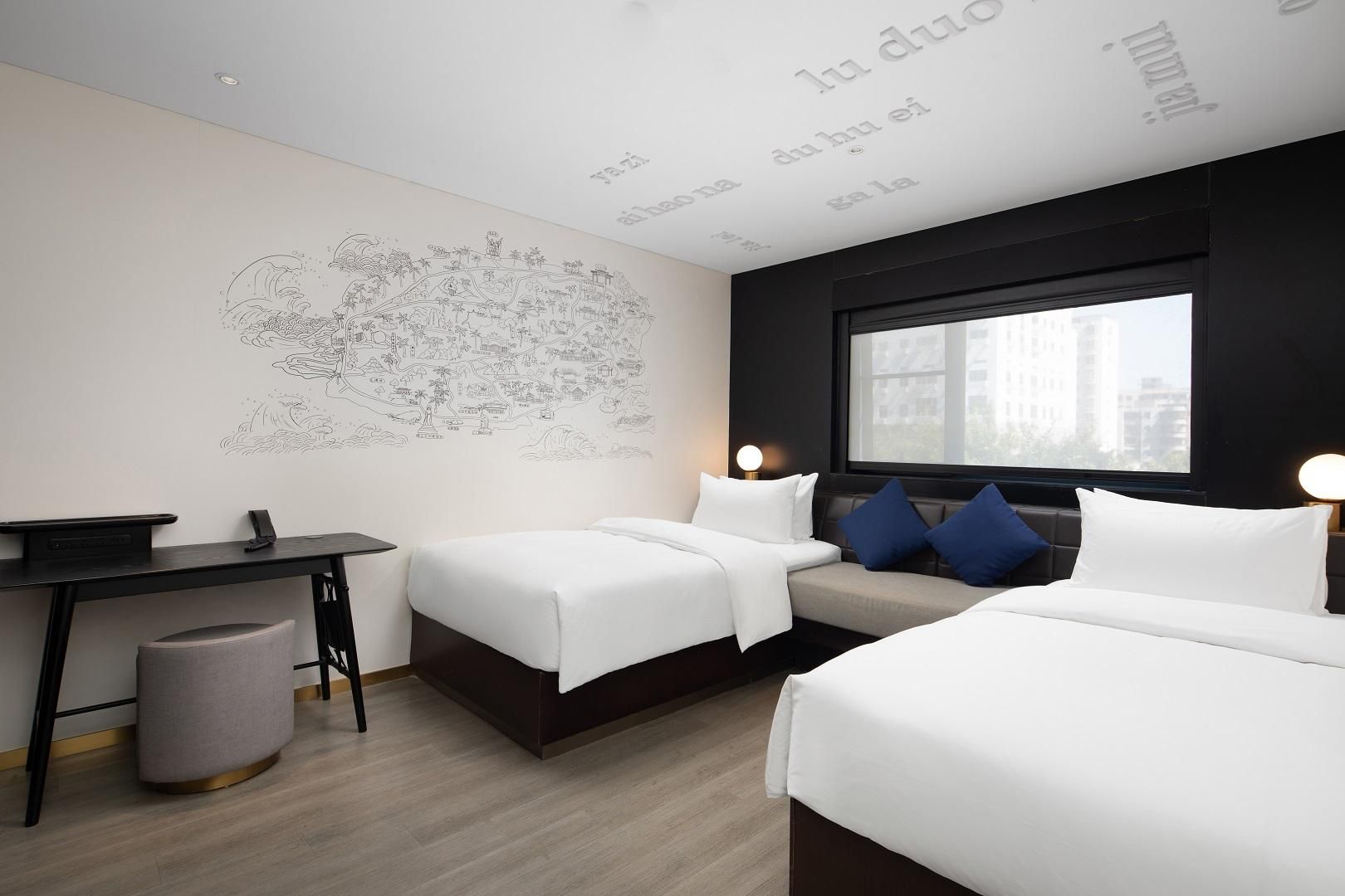 A hotel room with two beds, a desk, a chair, a trash bin, and a large window.
