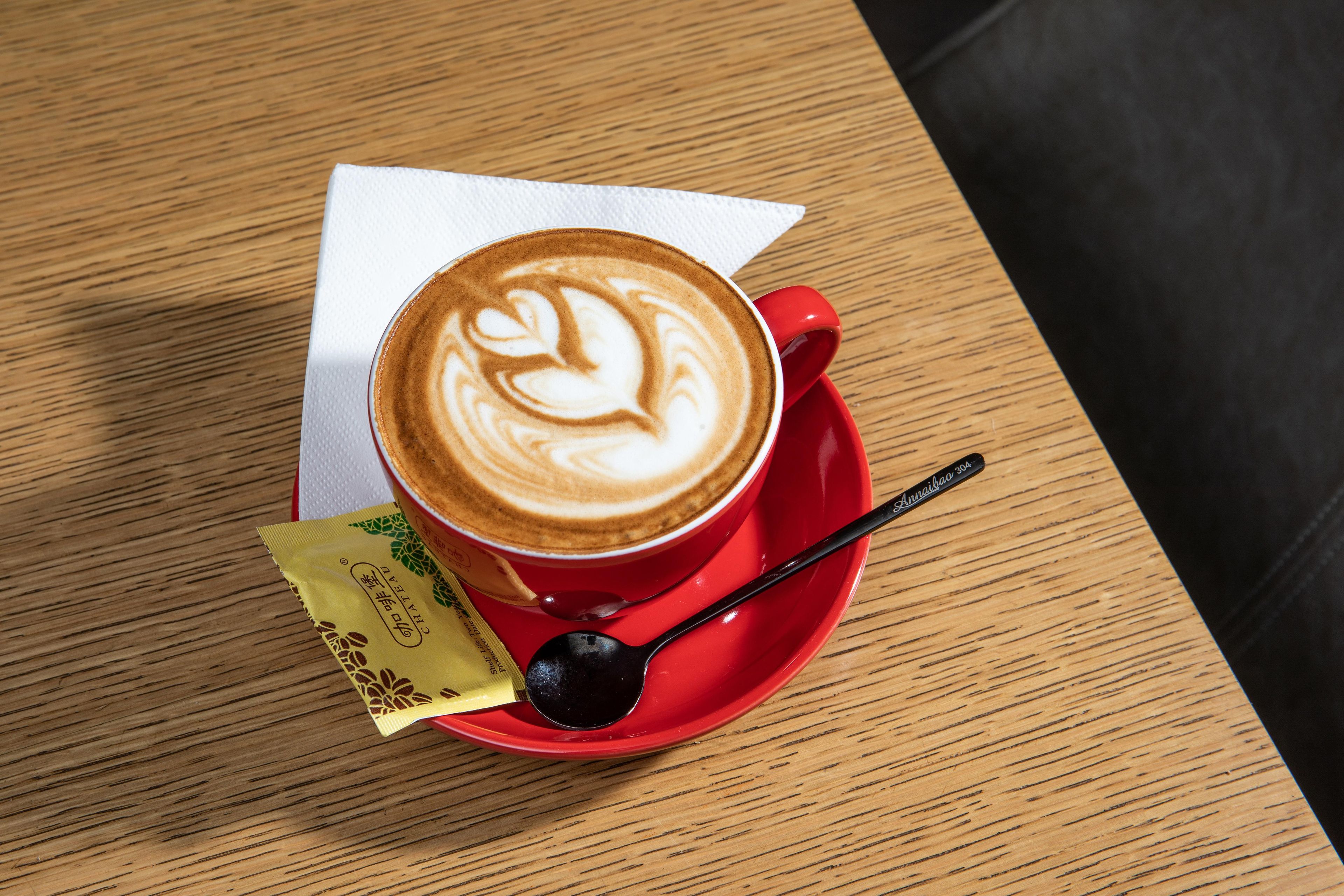 A cup of coffee with latte art on a red saucer, a spoon, and a packet of creamer on a wooden table.