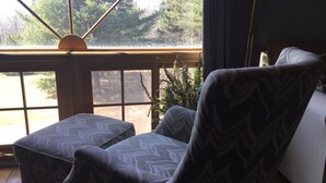 Una habitación acogedora con un ventanal grande, un sillón estampado, un reposapiés pequeño y una lámpara.