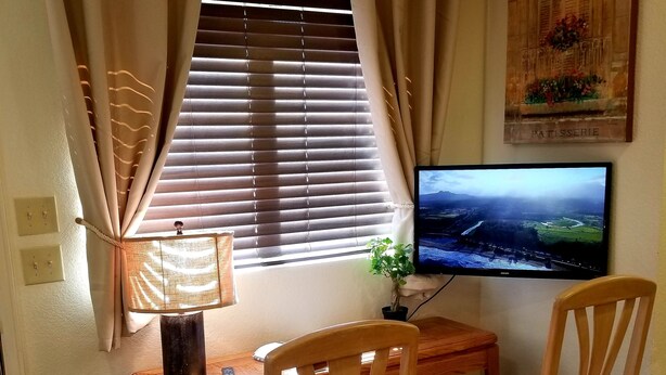 Ein Zimmer mit Schreibtisch aus Holz, zwei Holzstühlen, einem Fernseher, einem Fenster mit Jalousien und einem Wandbild.