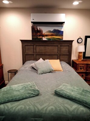 Une chambre à coucher avec un lit, des oreillers, une table de chevet, une lampe, un climatiseur et une horloge murale.