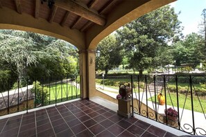 Een overdekt terras met betegelde vloer, ijzeren hekwerk en uitzicht op een aangelegde tuin.