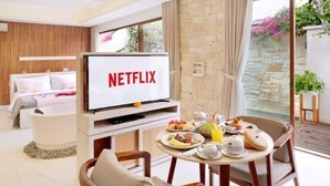 A modern hotel room with a bed, a TV displaying 'NETFLIX', a dining area with breakfast, and a view of a garden. - Glamour 1BR Private Villa Kuta (Bali)