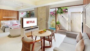A modern living room with a flat-screen TV displaying 'NETFLIX', a sofa, a dining table with chairs, and a view of a garden through a sliding door. - Glamour 1BR Private Villa Kuta (Bali)