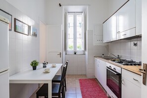Eine kompakte Küche mit weißen Schränken, einem Esstisch mit Stühlen, einem roten Teppich und einem Fenster mit Blick auf Gebäude.