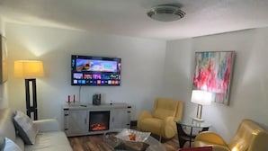 Een woonkamer met een bank, fauteuils, een glazen salontafel, een open haard, een televisie en een schilderij aan de muur.