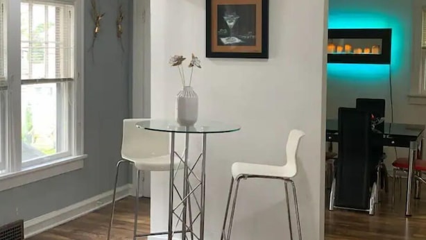 Een eethoek met een glazen tafel, twee witte stoelen, een vaas met bloemen en een lijst met een afbeelding aan de muur.