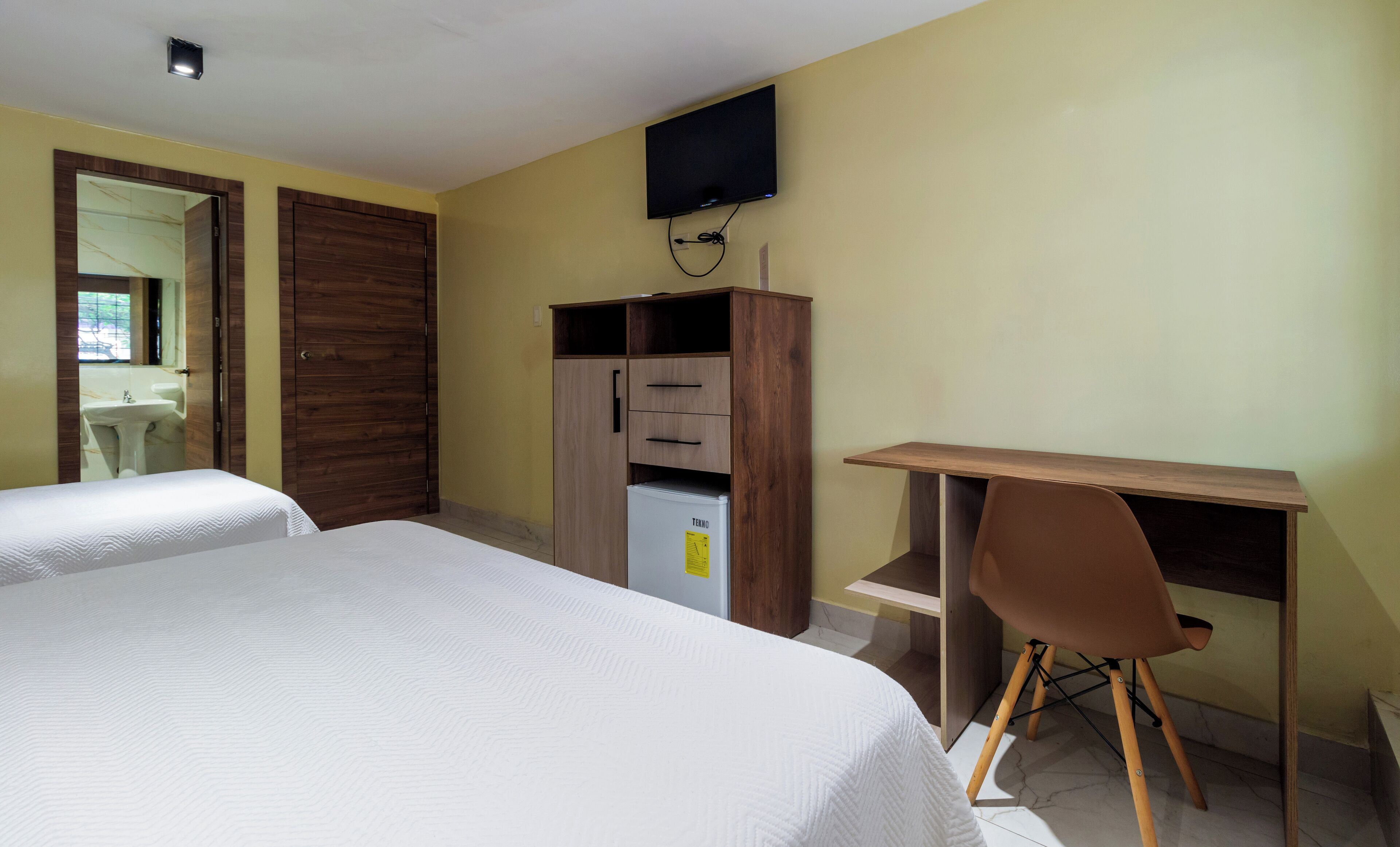 A bedroom with two beds, a wooden wardrobe, a desk with a chair, and a TV mounted on the wall.