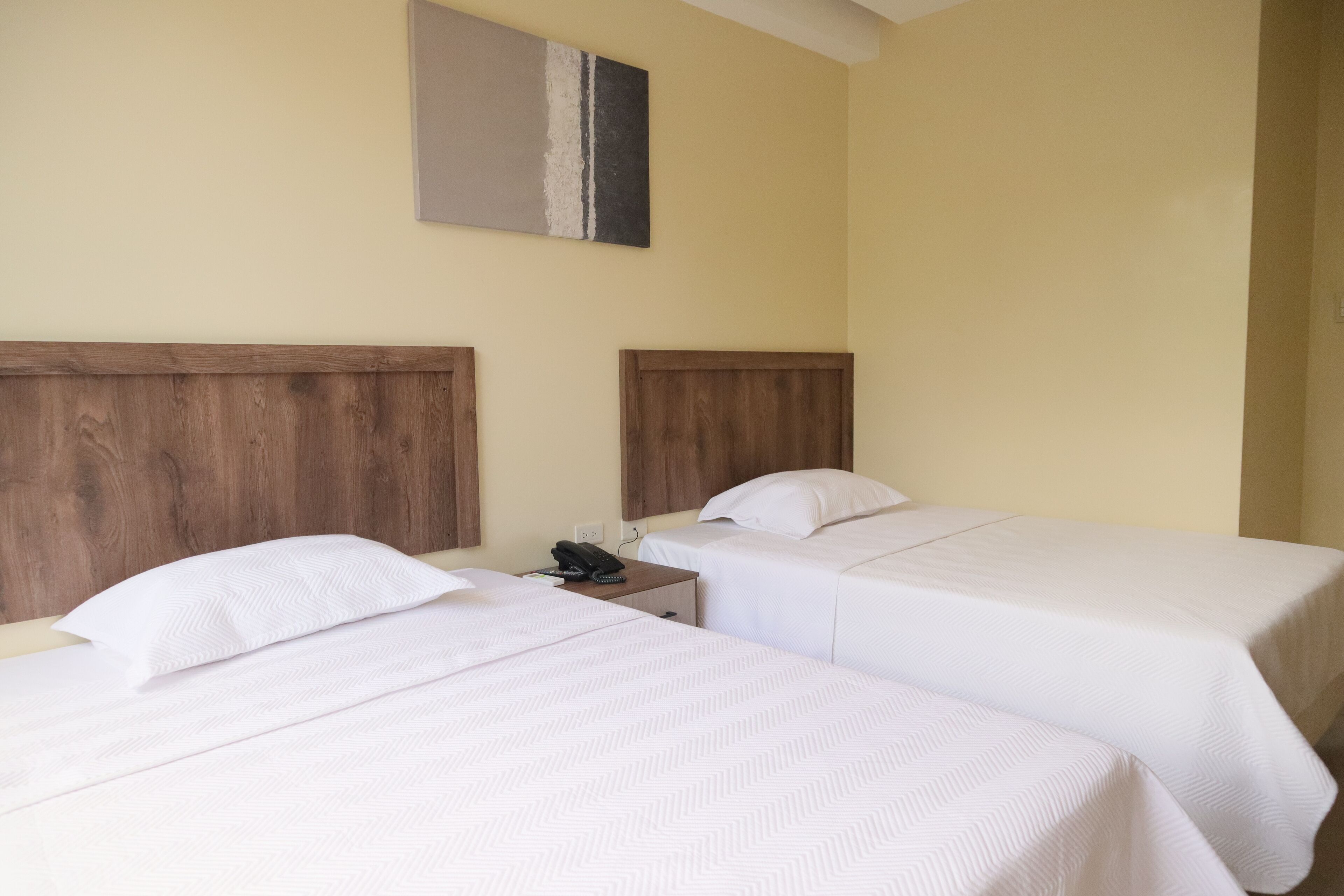 A hotel room with two single beds, wooden headboards, a nightstand with a phone, and a painting on the wall.