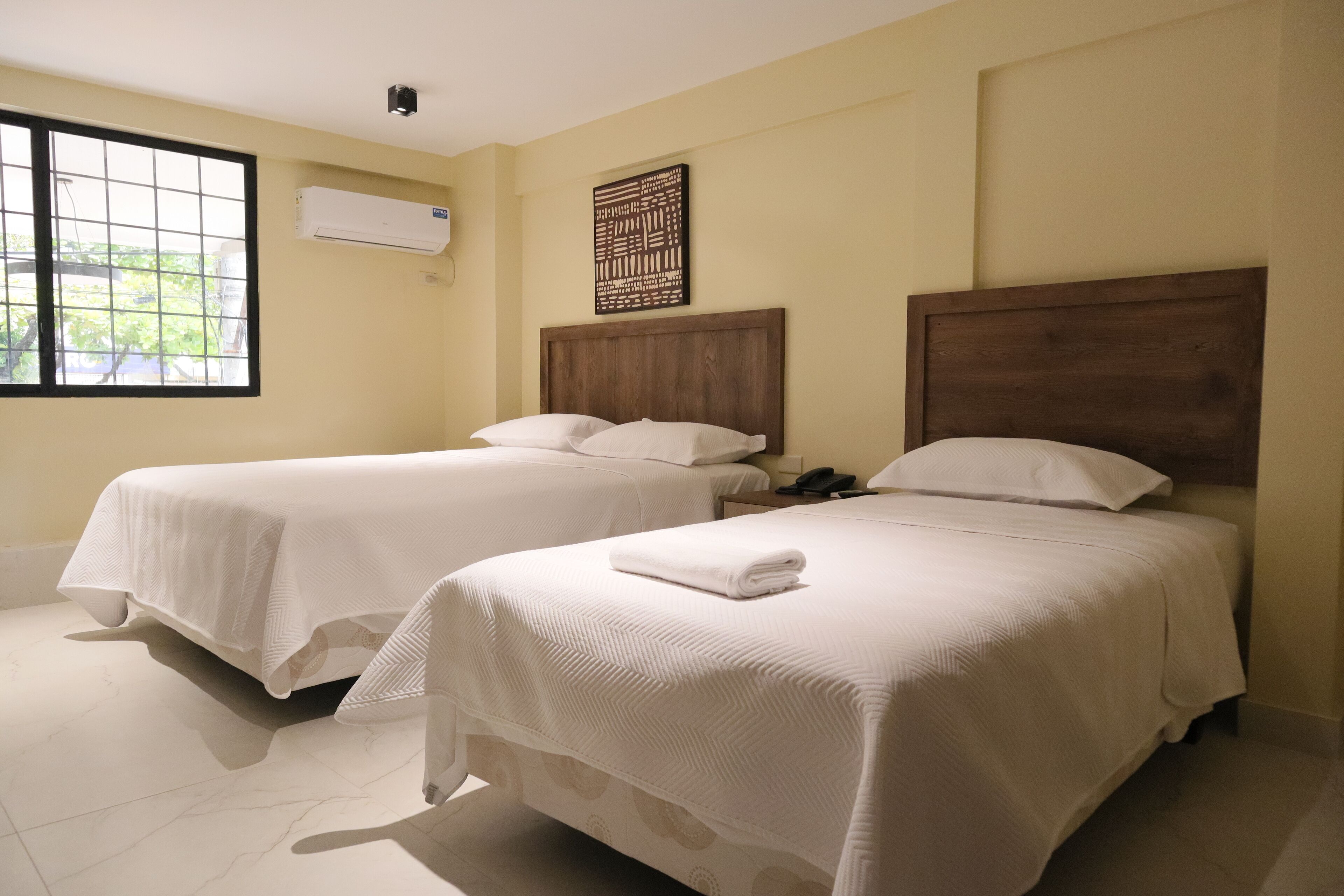 Two single beds with wooden headboards, a window with a view of greenery, and an air conditioning unit on the wall.