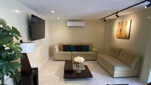 A modern living room with a beige sofa set, a coffee table with a vase of flowers, a black armchair, and a flat-screen TV mounted on the wall. - Hotel Sada (Guayaquil)