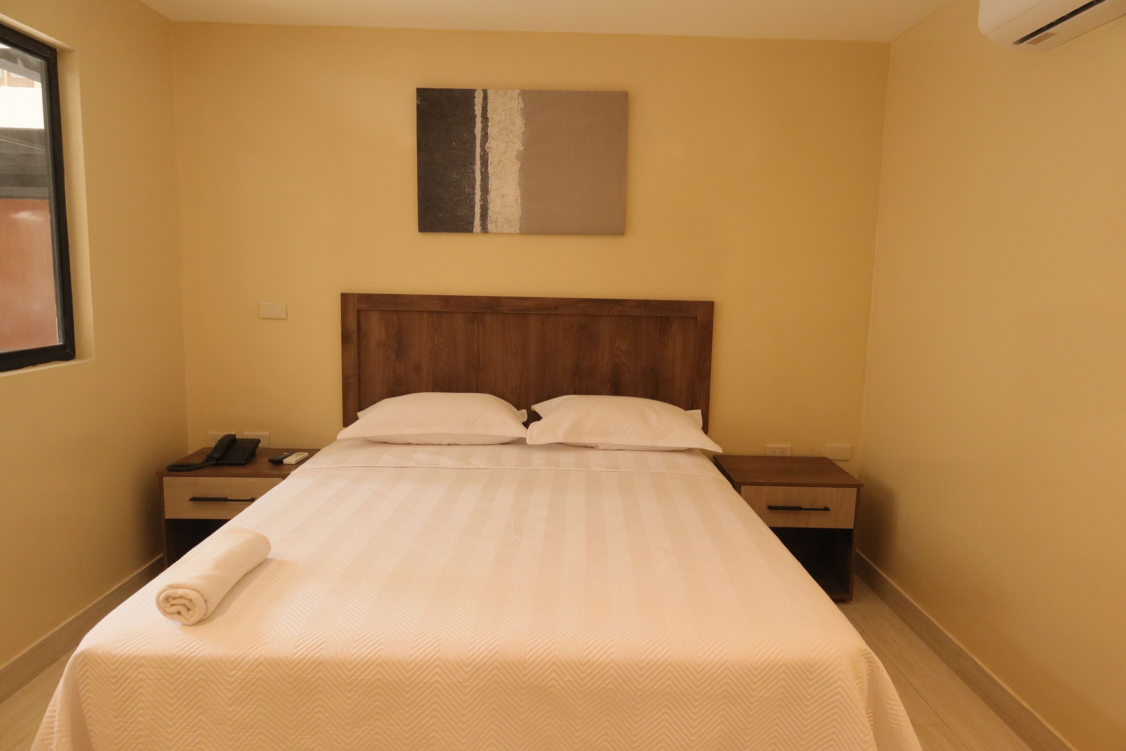 A hotel room with a wooden headboard, a bed with white linens, a bedside table with a phone, and a painting on the wall.
