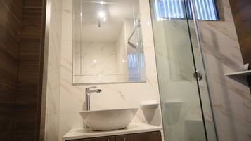 Un baño con ducha envuelta en vidrio, un lavabo blanco tipo recipiente y un espejo sobre el lavabo.
